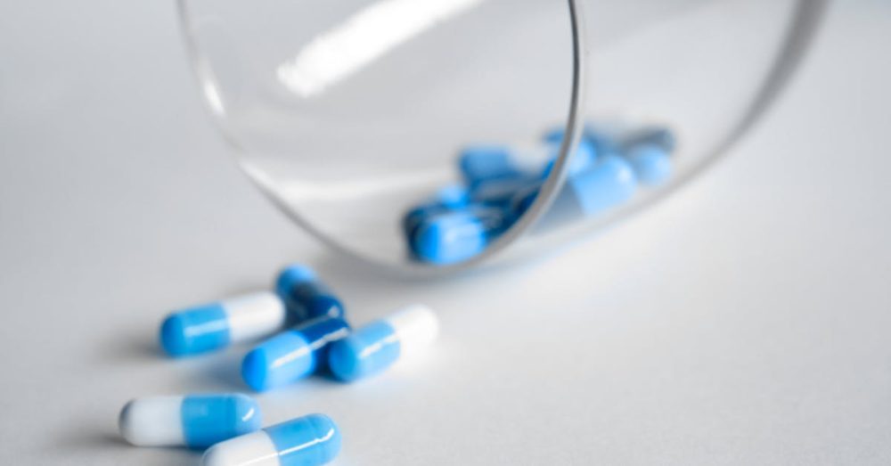 A close-up of blue and white capsules spilling from a glass on a light background.