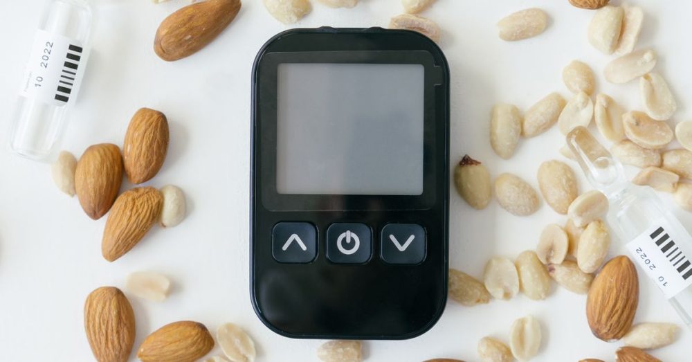 A top view of a glucose meter surrounded by almonds, syringes, and peanuts on a white background.