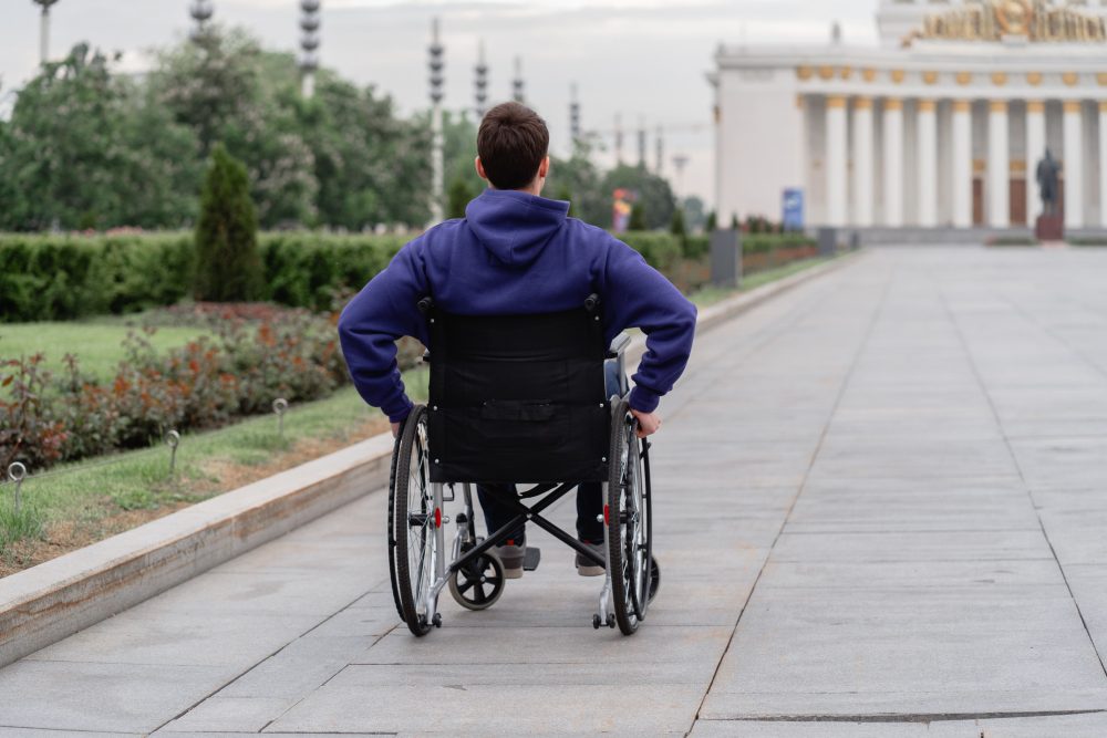 Man in a wheelchair wearing a blue jacket, seen from behind in a public park area, creating an inclusive atmosphere.