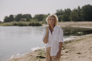Elderly woman in a white shirt standing by a tranquil lake on a sunny day.